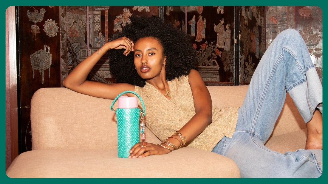 Girl with back fro and gold vest in jeans sitting on gold couch with a green mermaid loomi water bottle 